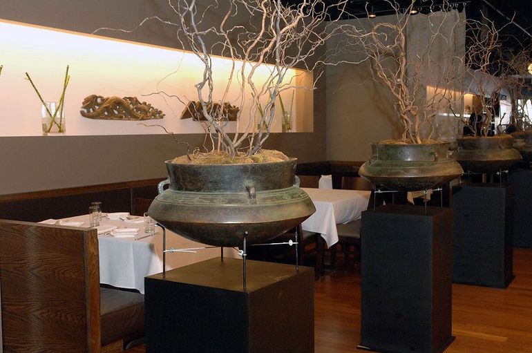 Restaurant interior with large bronze pots holding bare branches on black pedestals. Tables set with white tablecloths.
