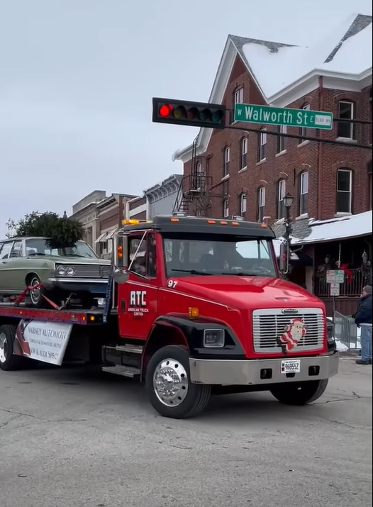Red tow truck carrying a classic car on a snowy street, under a traffic light.