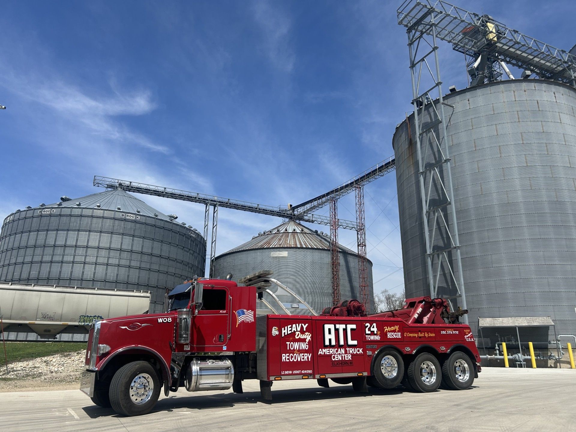 Red tow truck parked near large grain silos under a blue sky.