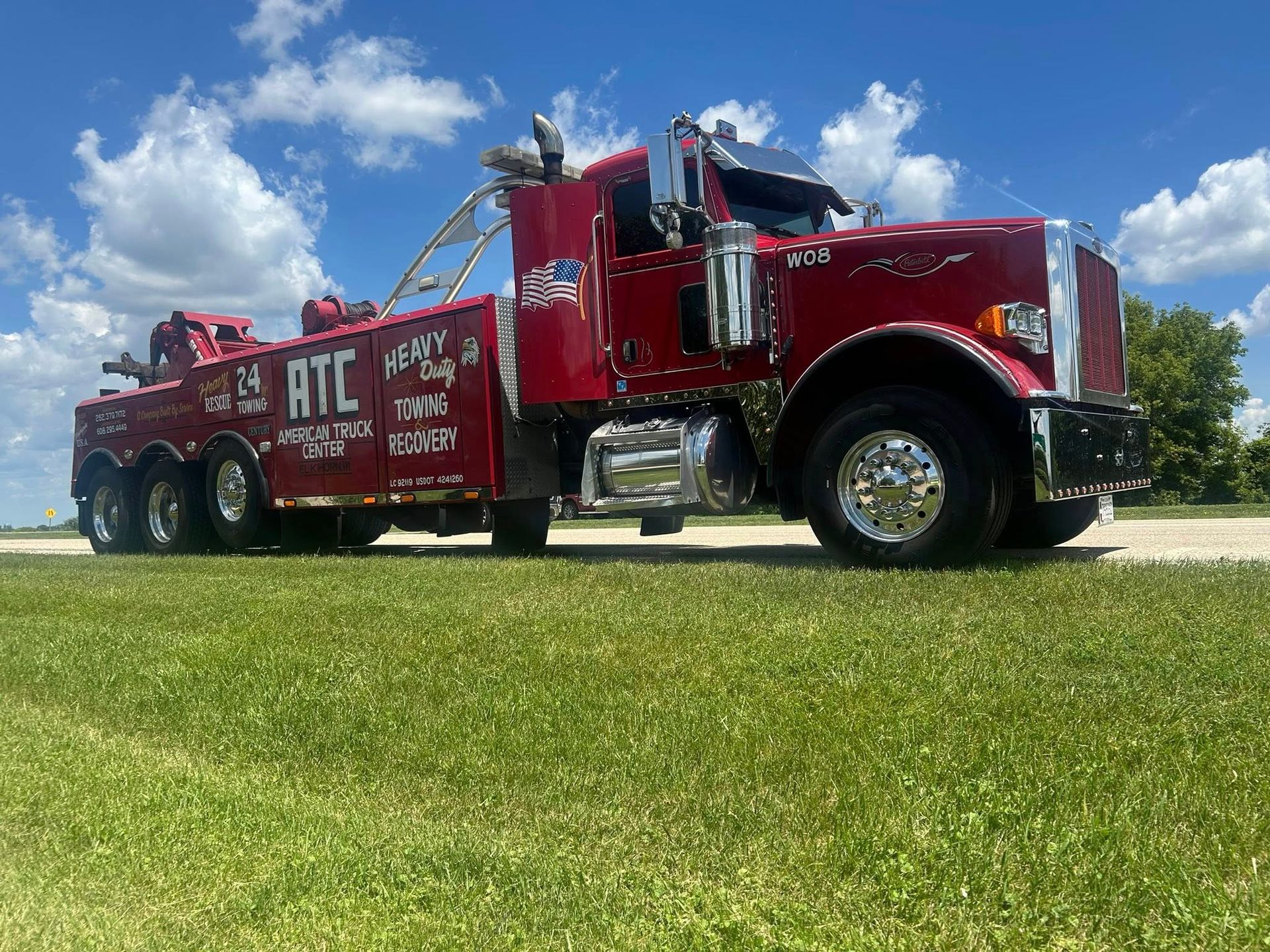 Red tow truck with chrome accents on a grassy field under a blue sky with clouds.