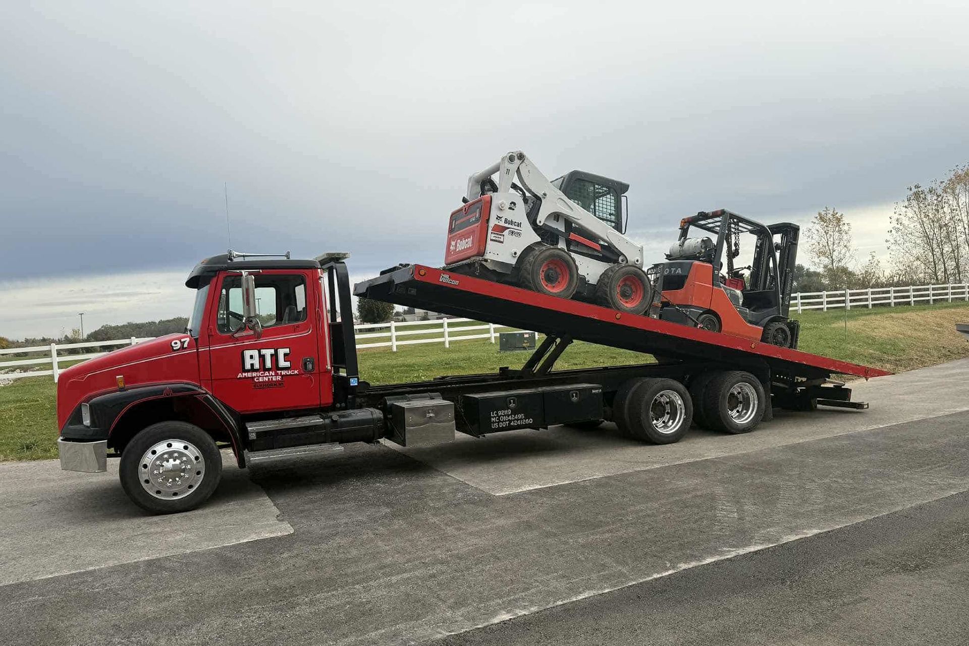 Red tow truck transporting a skid steer loader and a forklift on a flatbed trailer. Cloudy day.