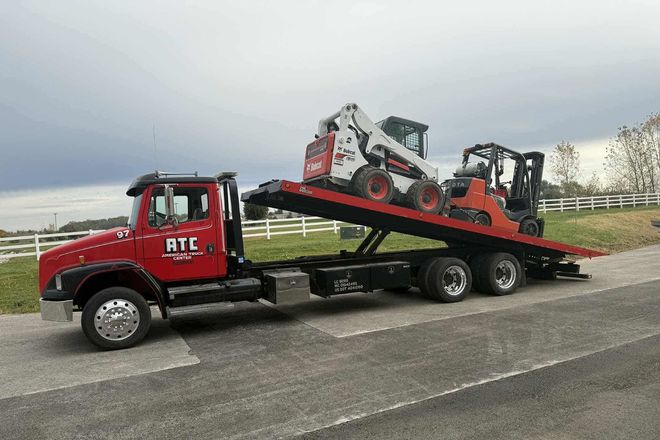 Red tow truck transporting a skid steer loader and a forklift on a flatbed trailer. Cloudy day.
