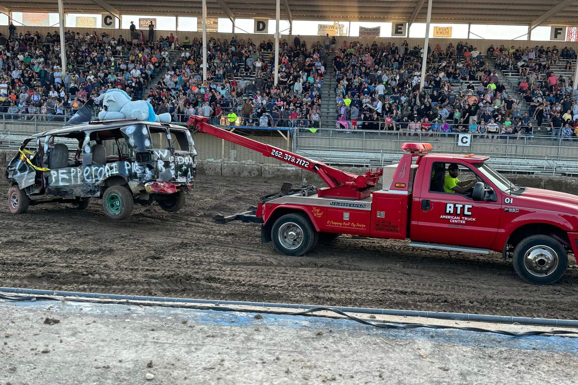 A red tow truck pulls a wrecked black SUV from a muddy arena during a demolition derby. Large crowd watches.