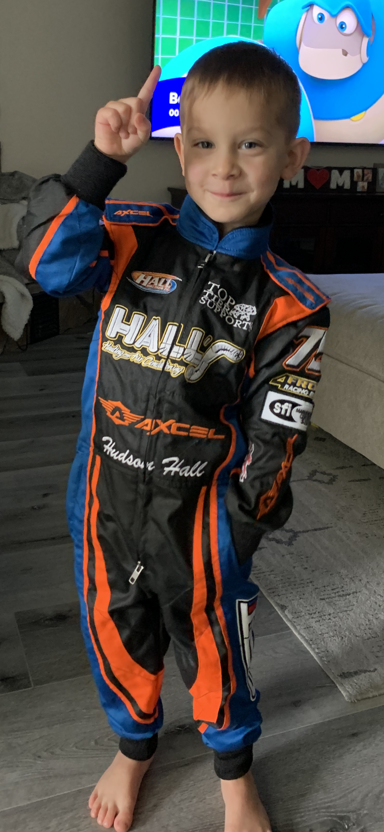 A young boy wearing a racing suit is standing in a living room