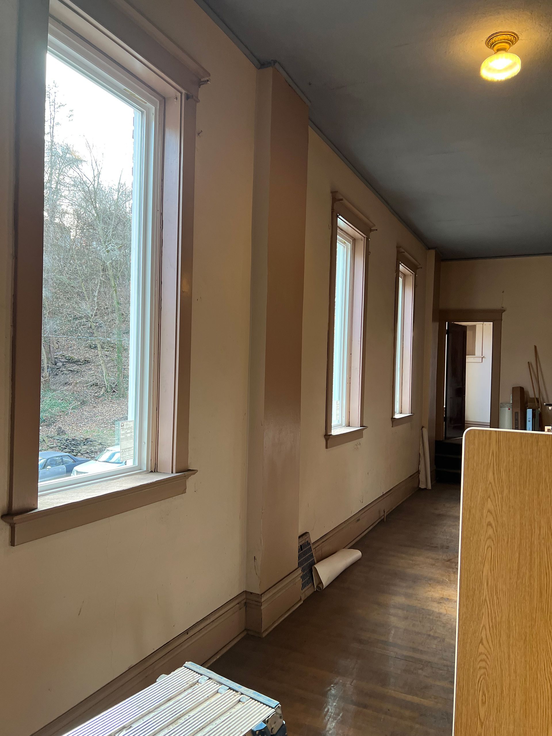 Narrow room with three windows, curtains, and a doorway. Cream walls, wood floors, and a light fixture.