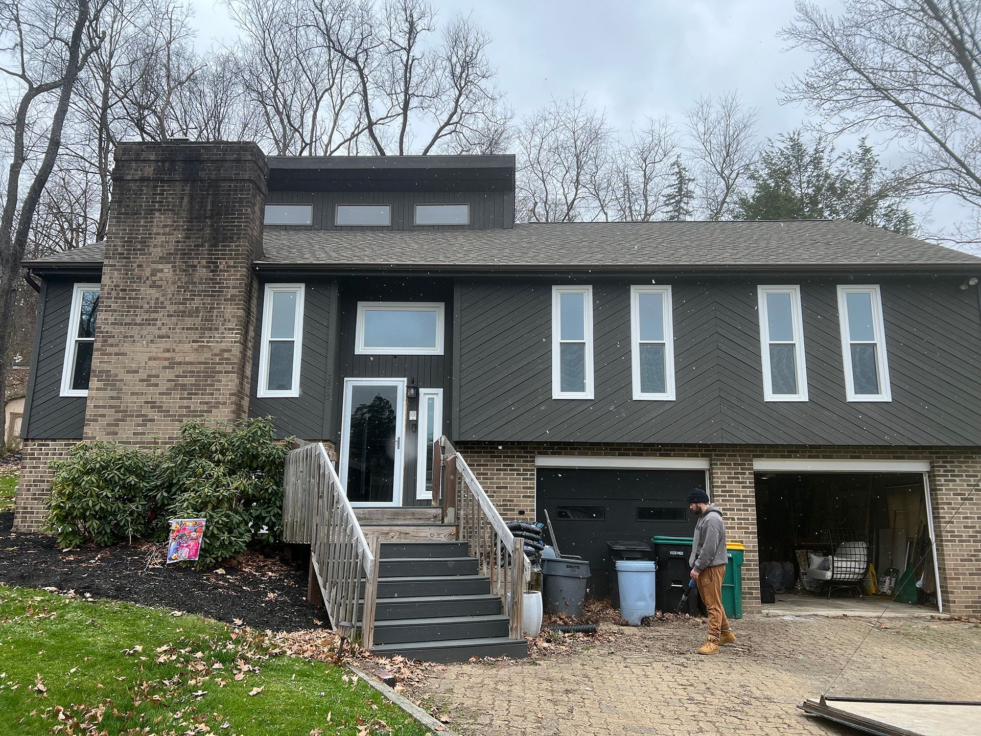 Two-story house with gray siding, white-trimmed windows, and a stone chimney. Person stands near the garage.