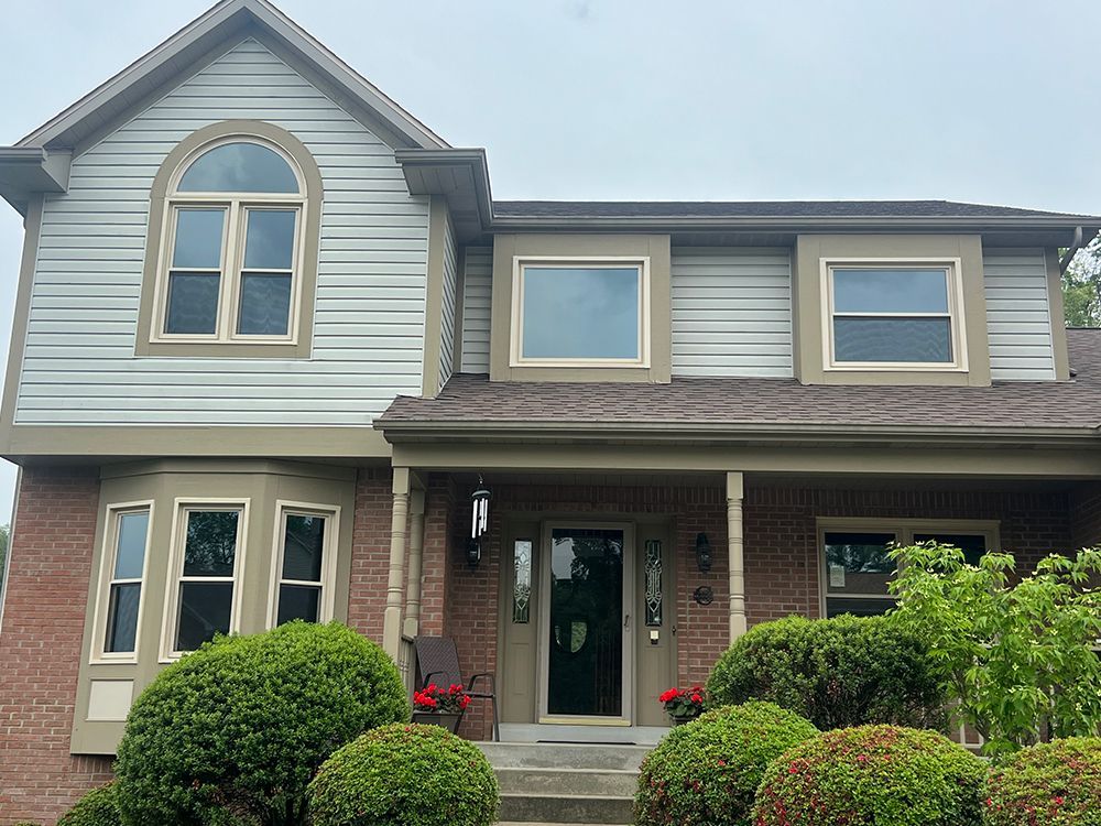 Two-story house with red brick, tan trim, and light blue siding, surrounded by green bushes and a cloudy sky.