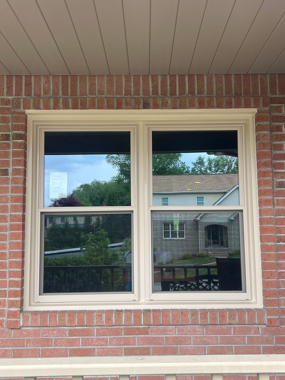 Window in brick wall, reflecting trees and a house, beige trim.