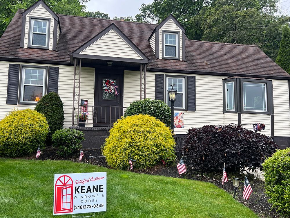 House with cream siding, dark roof, bushes, American flags, and a Keane sign on a slight hill.