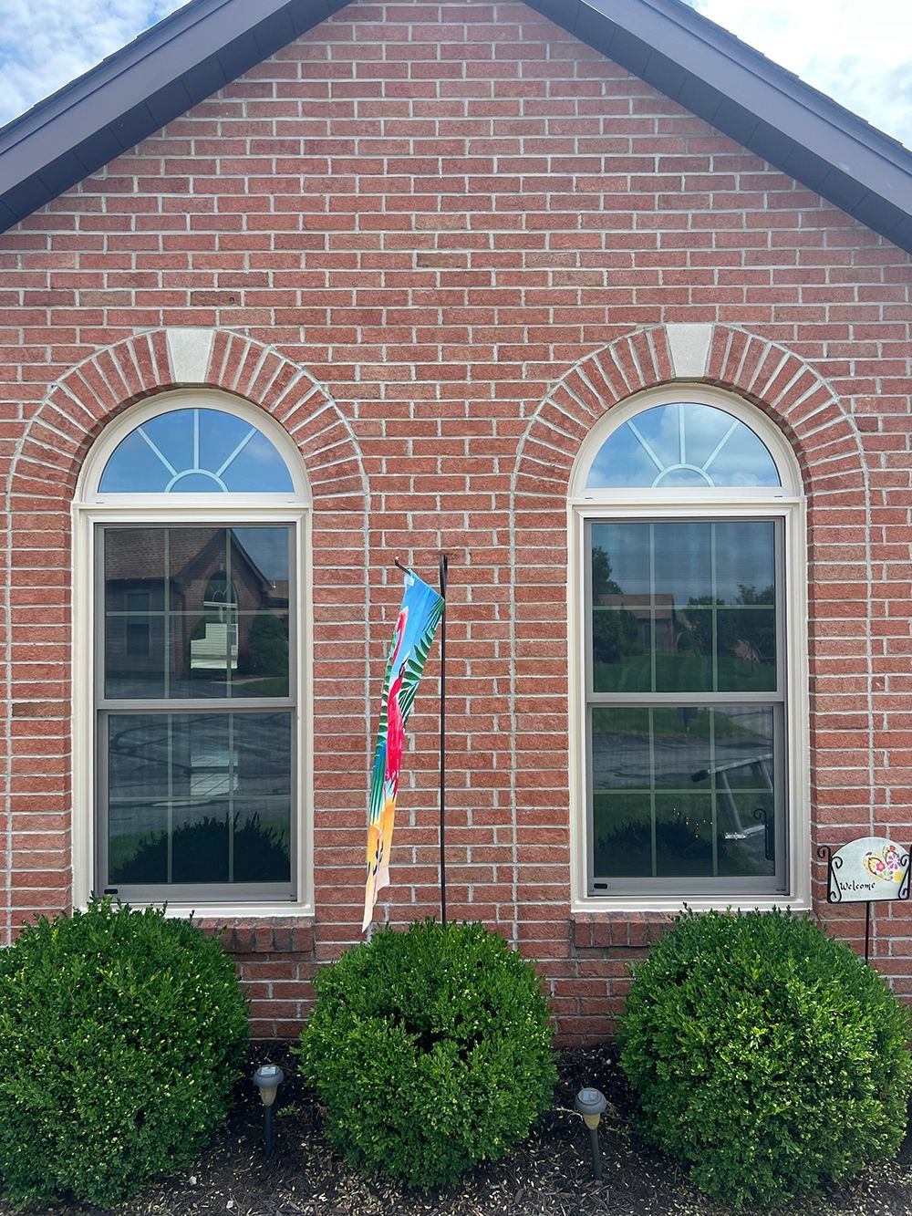Two arched windows in a brick house with green bushes beneath, and a decorative flag in front.