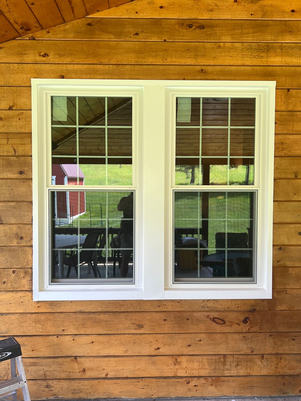Two white-framed windows with grilles in a rustic wooden wall. Reflections show a green yard, and a red building.