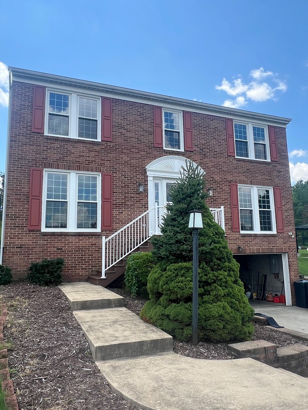 Red brick house with white trim, shutters, and entrance stairs, under a blue sky.