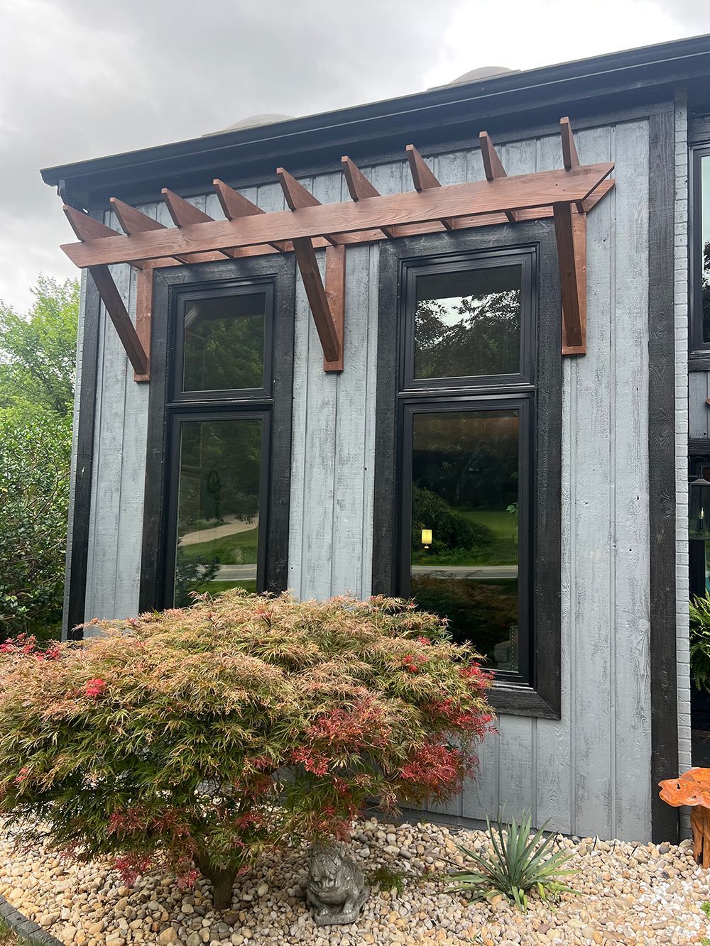 Gray house with dark windows and a brown pergola; a small tree in front.