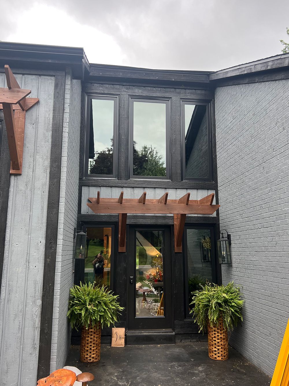 Gray building entrance with dark wooden door, windows, and pergola. Ferns in baskets flank the door.