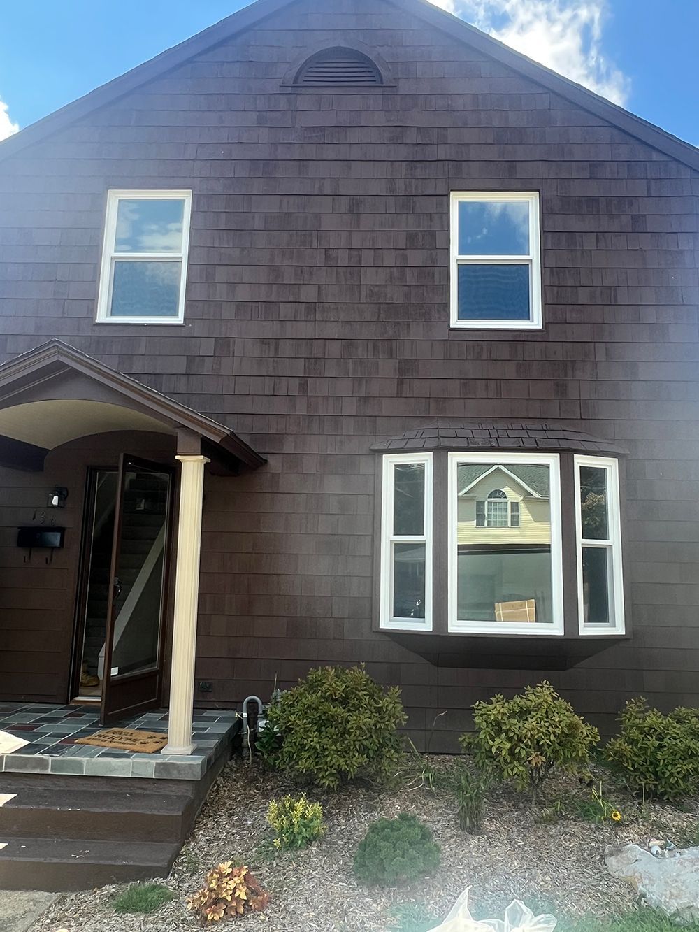 Brown two-story house with white-framed windows. The entrance has a porch and a door. The sky is blue.