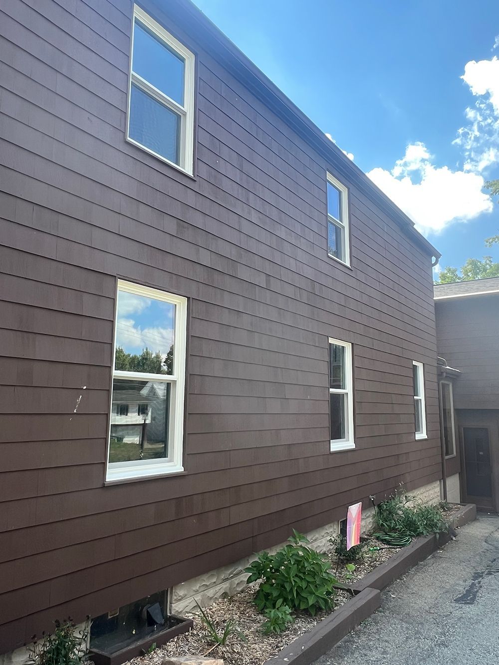 Brown-sided building with white-framed windows against a blue sky. Small garden along the bottom edge.