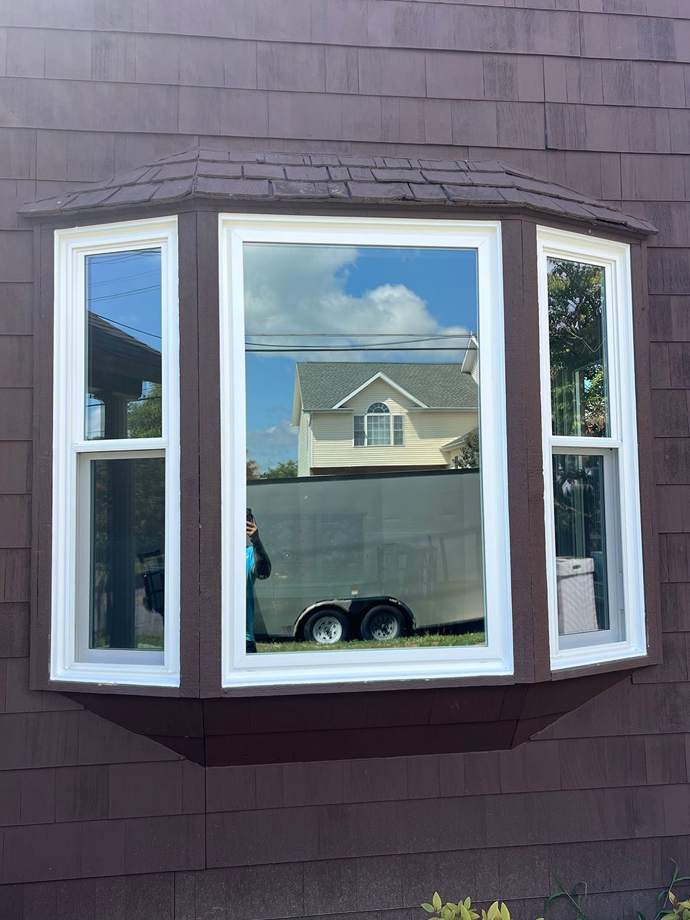 Bay window on brown shingled house reflects a house, trailer, and blue sky.
