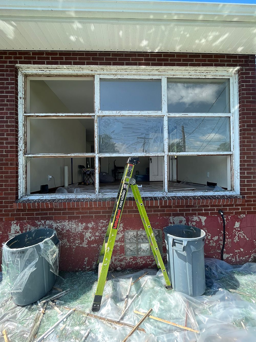 Window being replaced on a brick building, with a ladder, trash cans, and plastic sheeting.