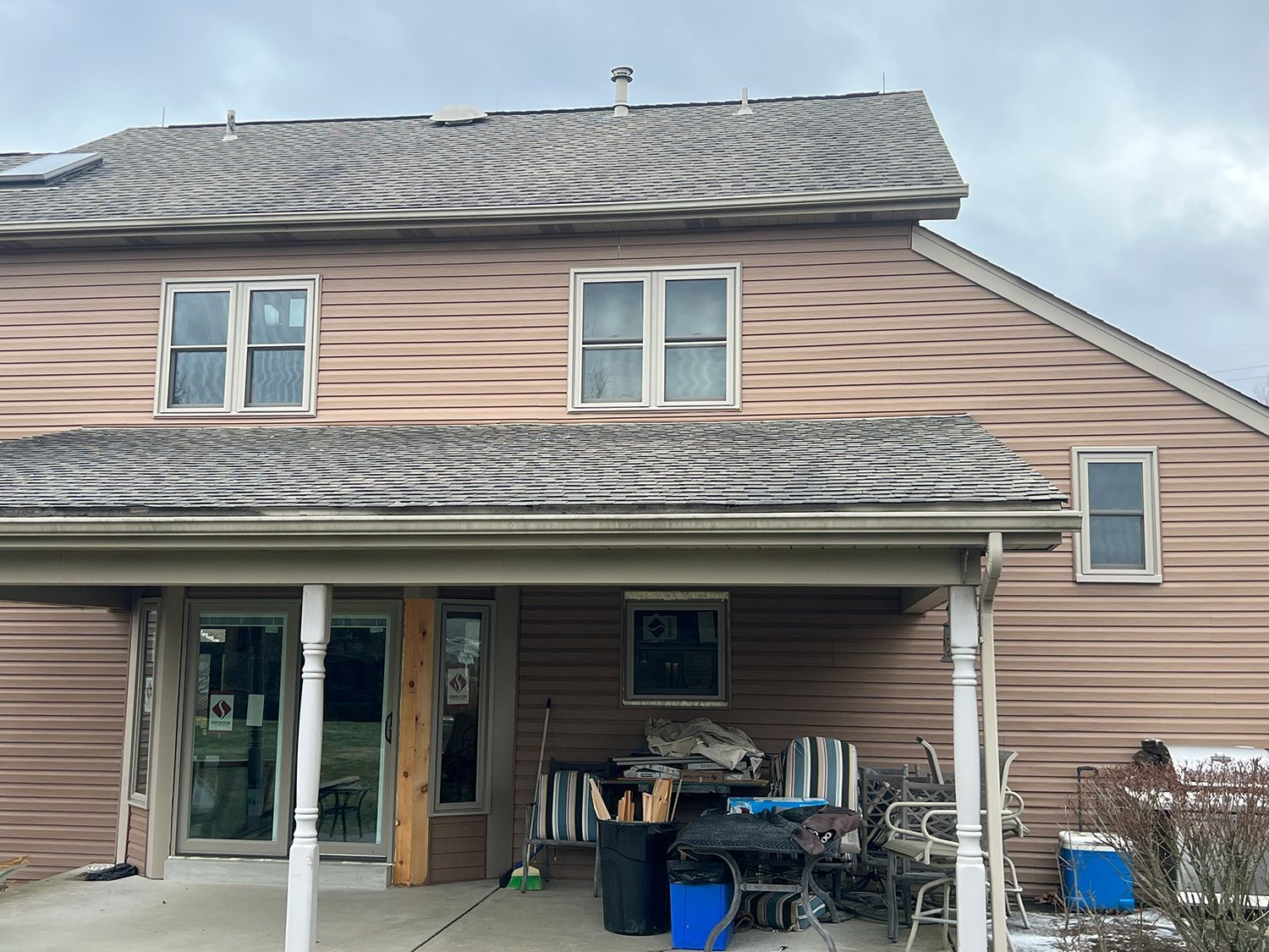 Beige siding house with snowy roof and porch, windows, and outdoor furniture. Overcast sky.