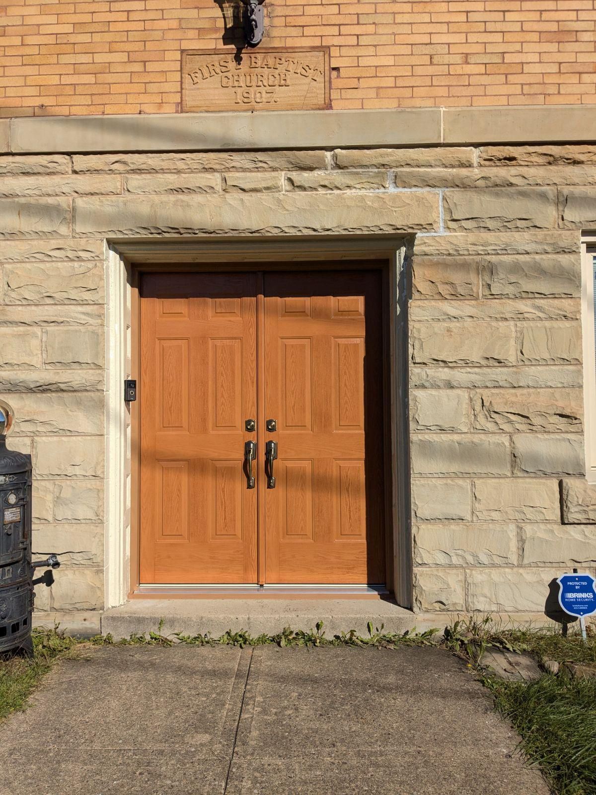 Double wooden doors with black handles in a stone building entrance, a concrete walkway in front.