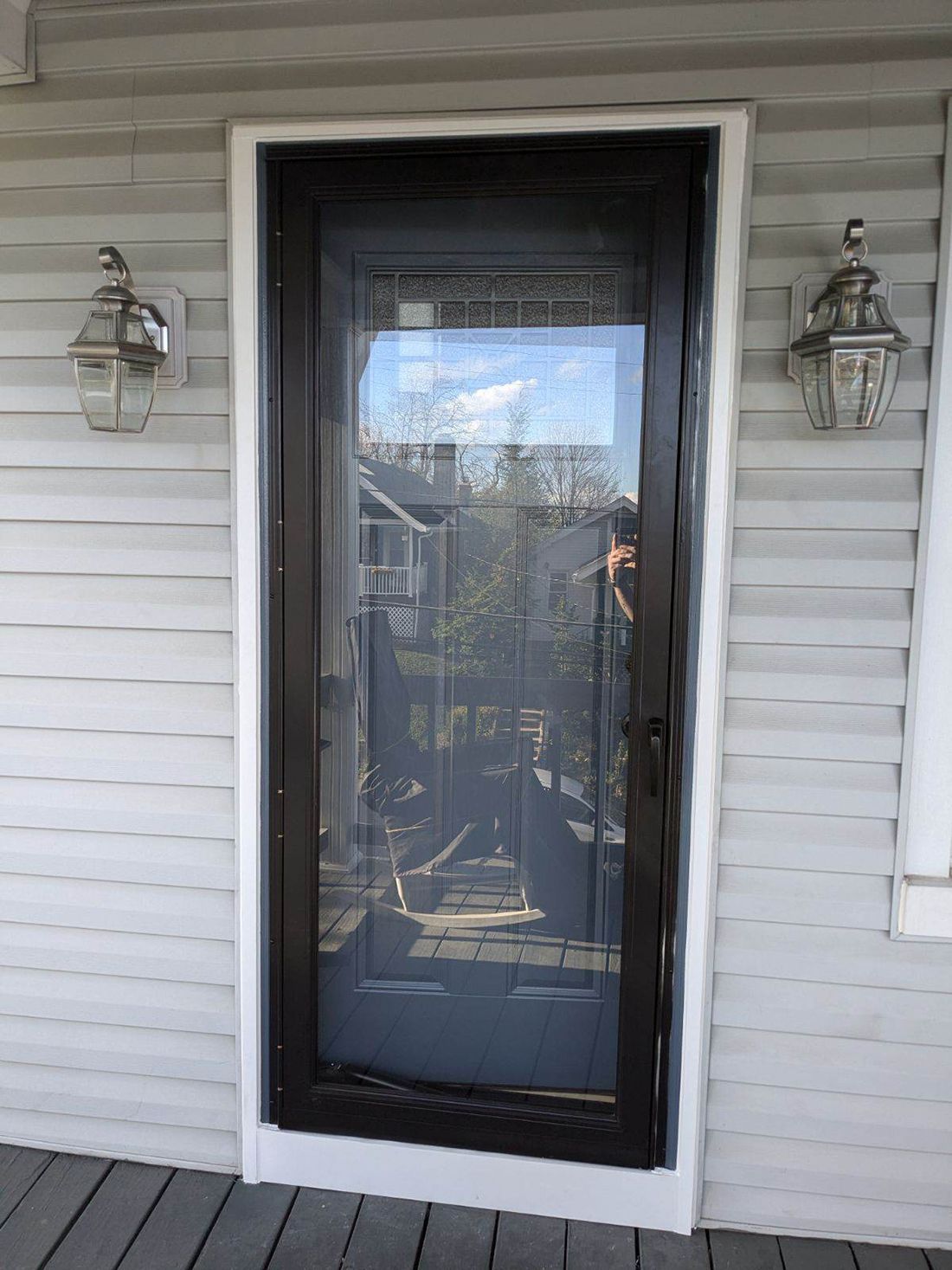 A dark-framed screen door with a glass panel is centered on a white building with light fixtures on each side.