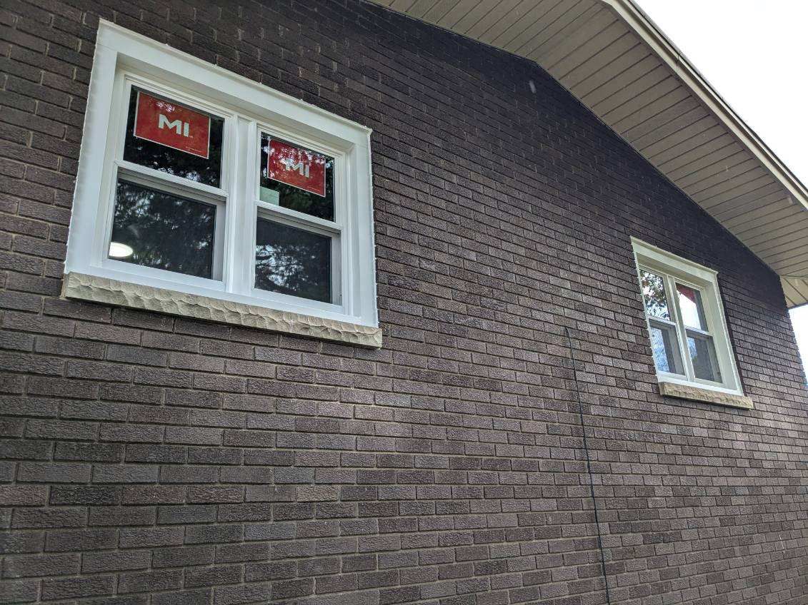 Brick house exterior with two white-framed windows. Dark brown brick and beige trim visible.