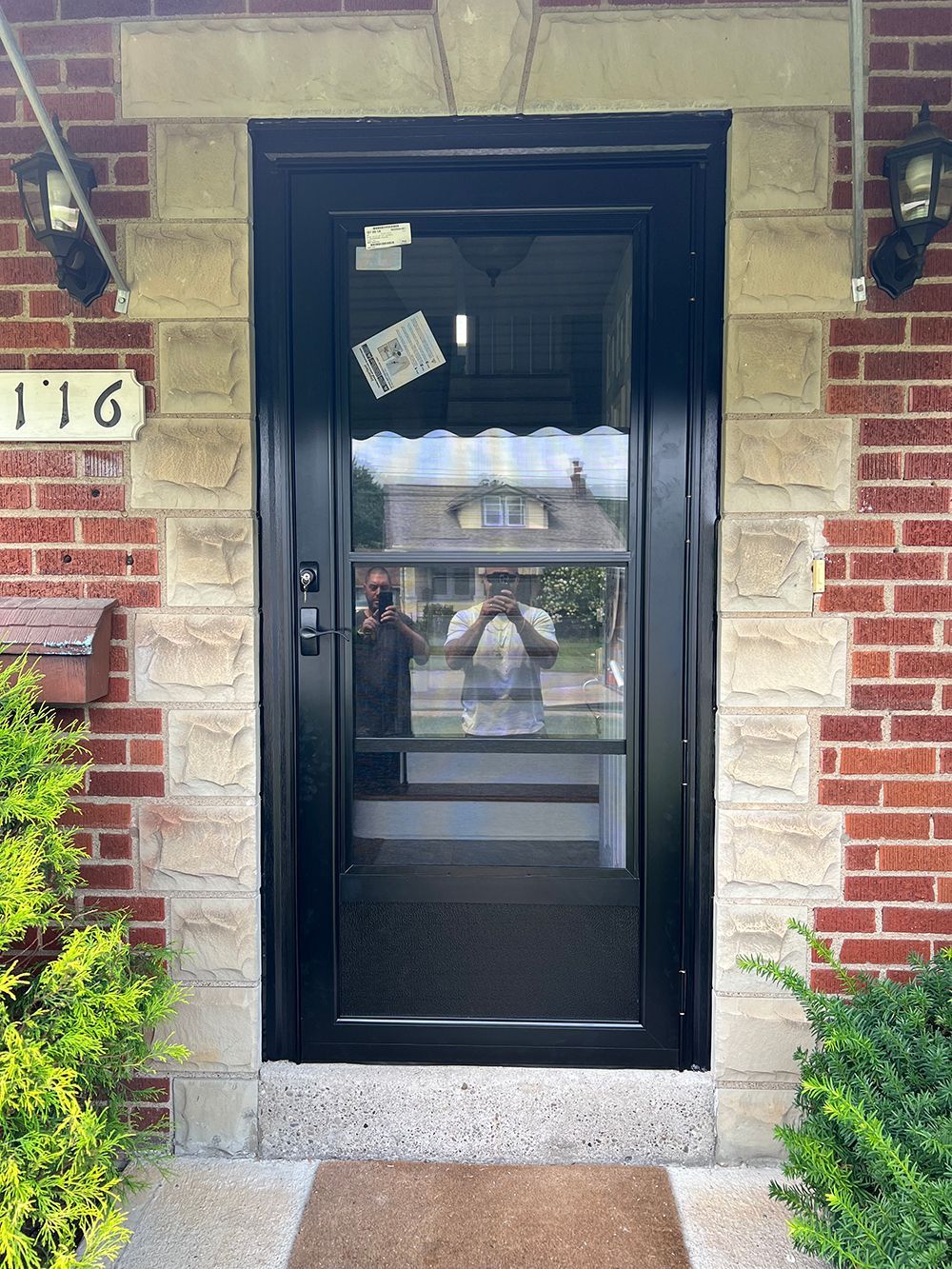Black screen door on a brick and stone house, with a person reflected in the glass.
