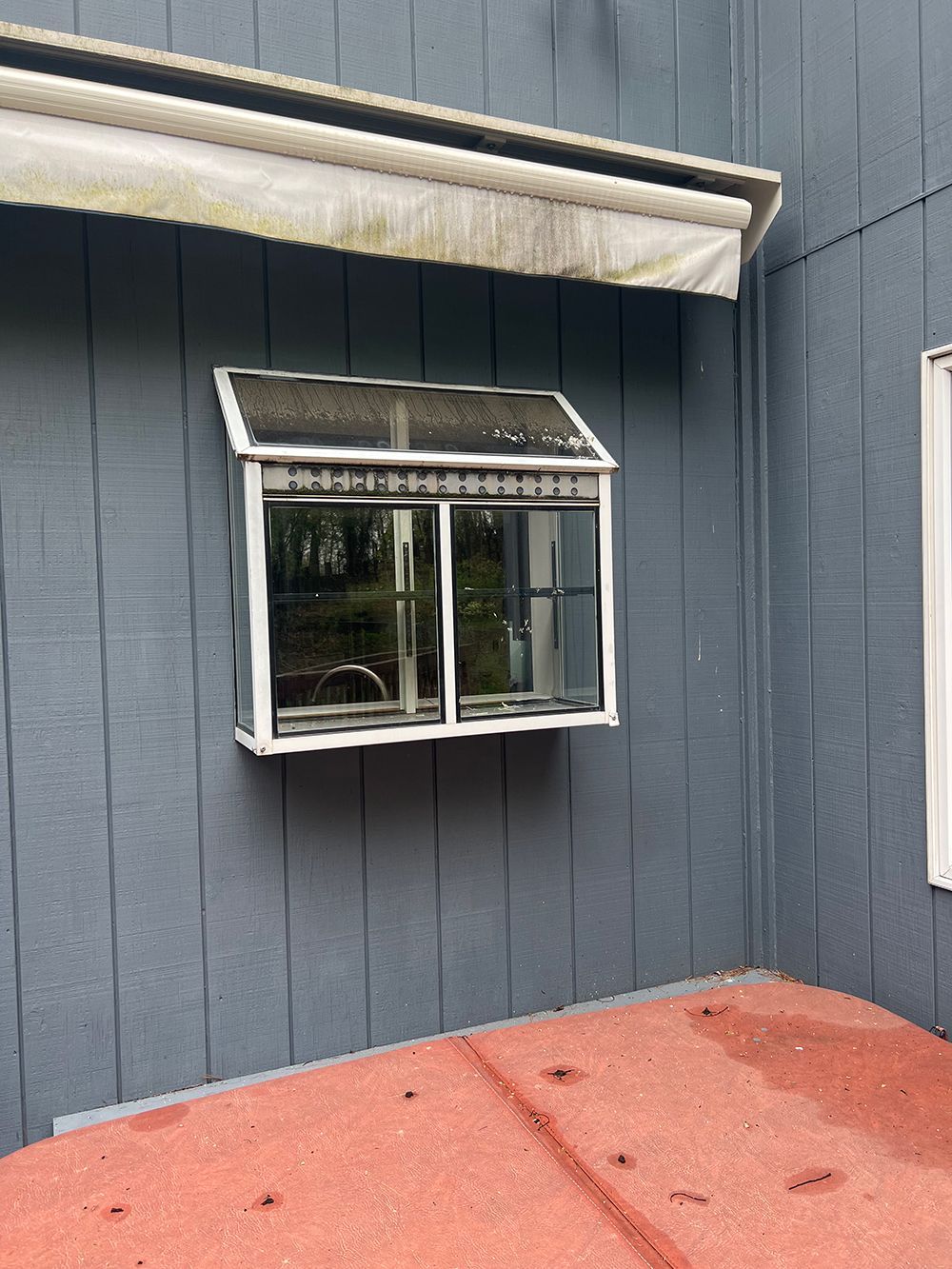 White-framed window on a gray-sided house. Red patio below. Overhanging awning is dirty.