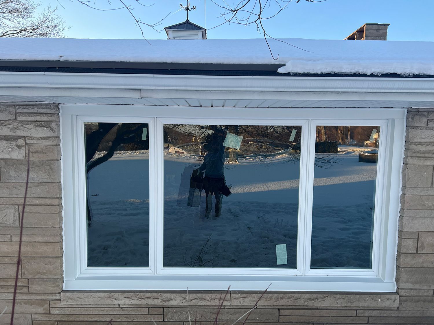White-framed window reflecting a snowy yard, person and dog; building with brick facade and snow-covered roof.