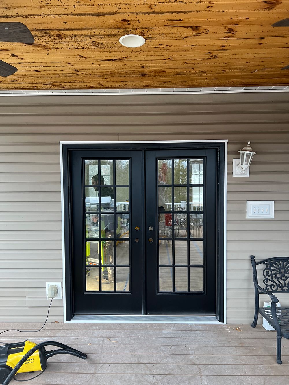 Black French doors with white trim, on a tan building, under a wood ceiling, with a bench nearby.