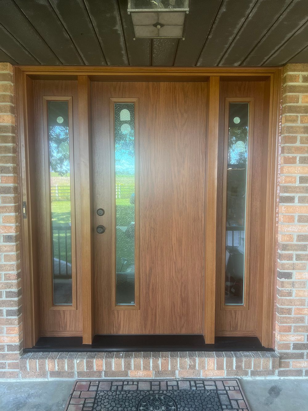 Wooden front door with glass panels, flanked by brick, under a light fixture.
