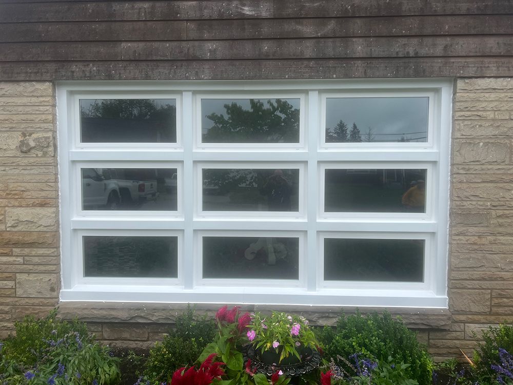 White-framed multi-pane window set in stone wall, reflecting trees, flowers in foreground.