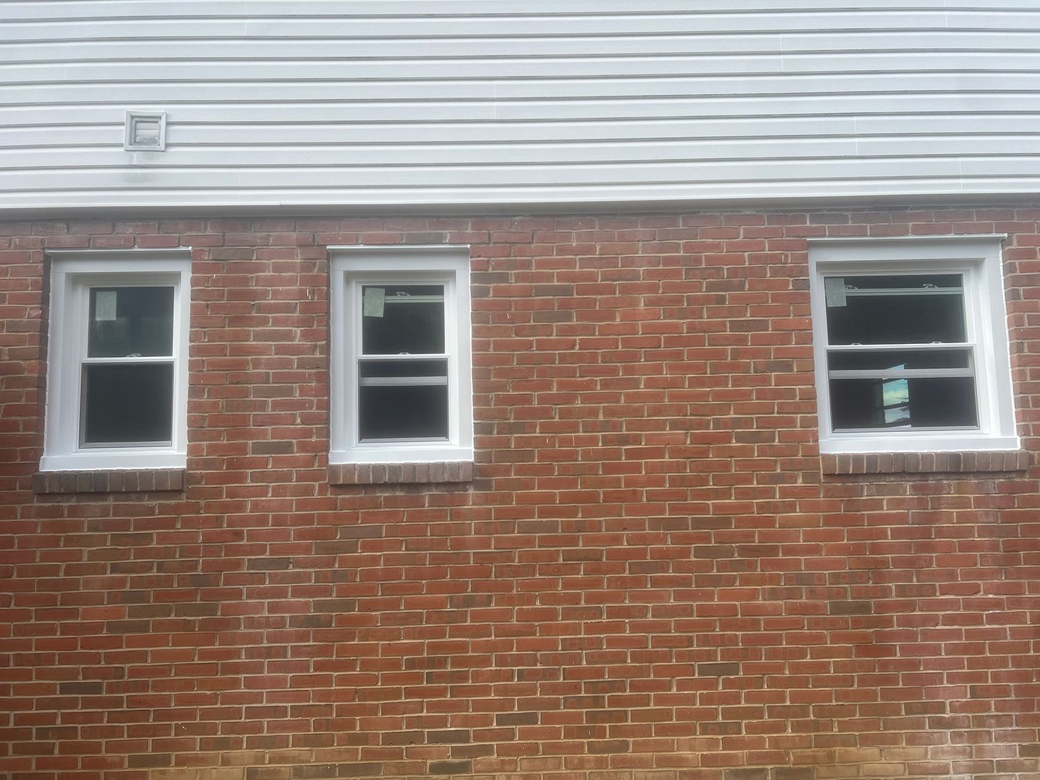 Three white-framed windows in a red brick wall beneath white siding.