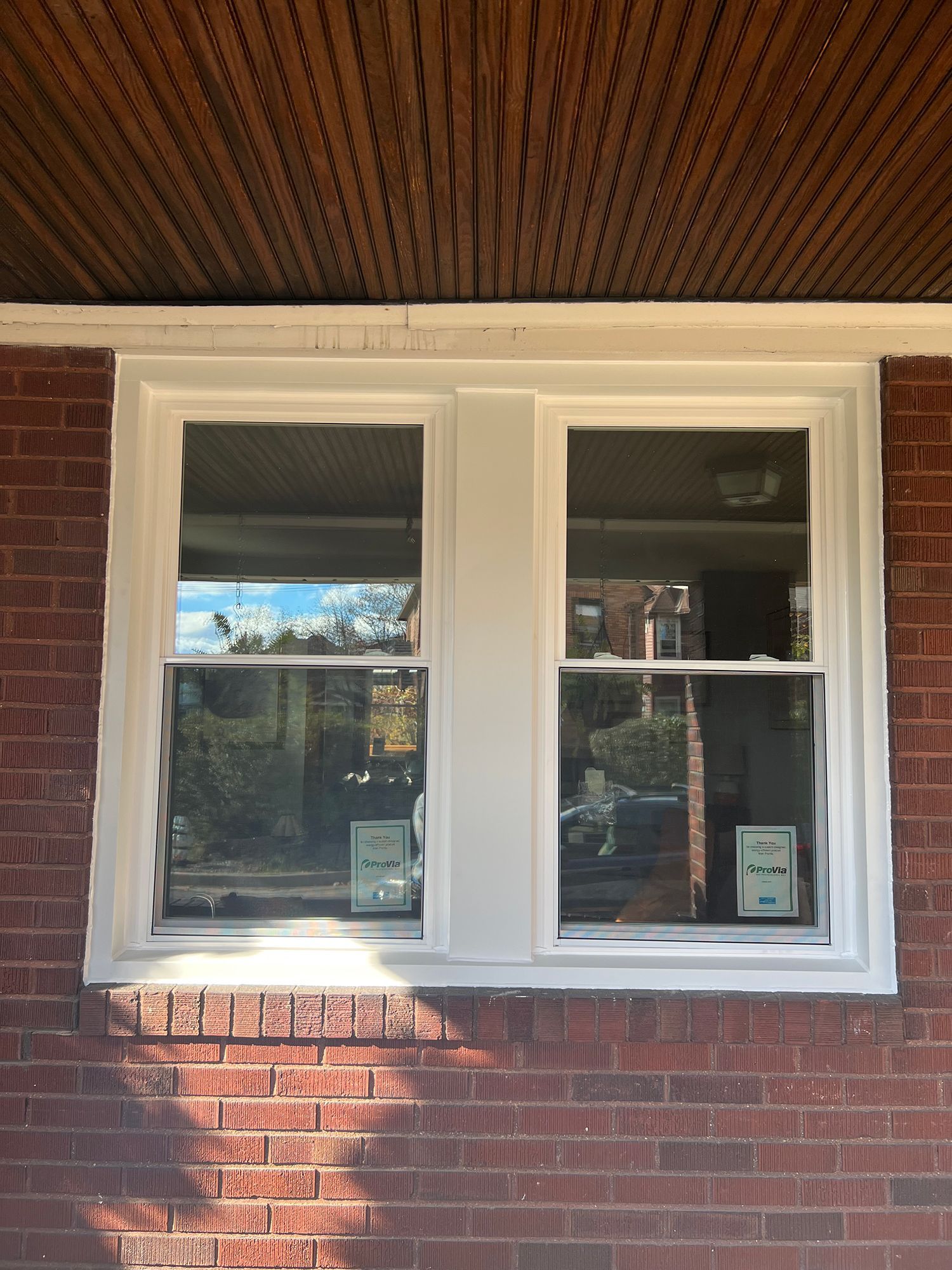 Two white-framed windows on a brick building, reflecting trees and a sunny sky.