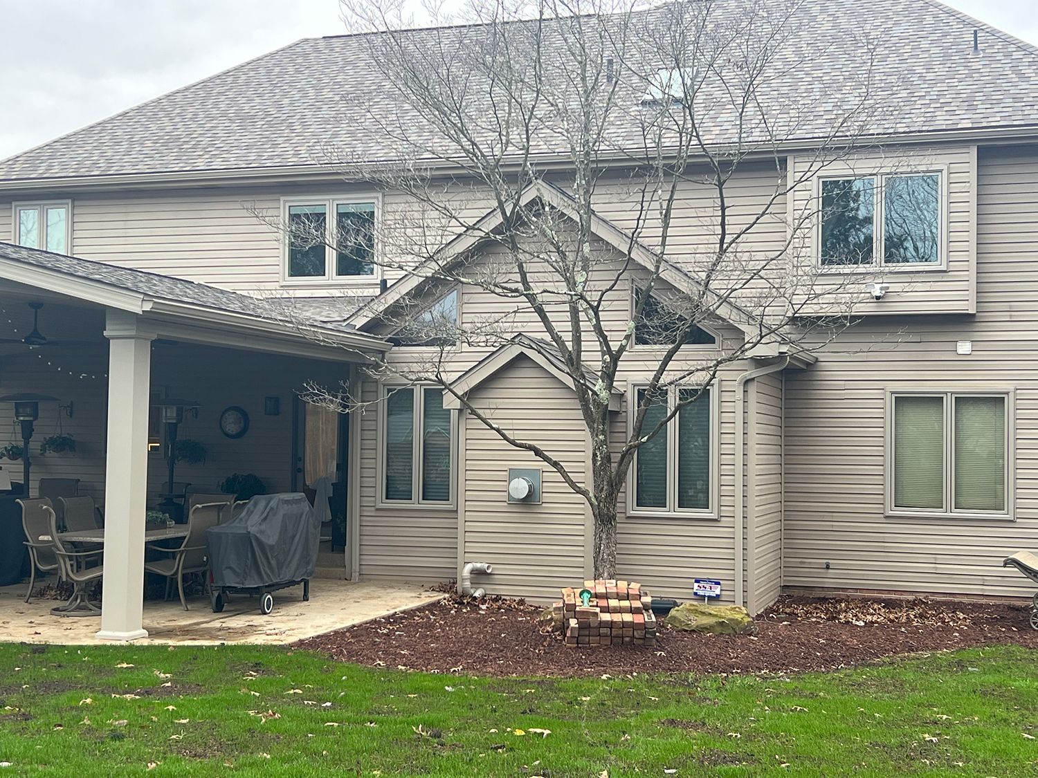 Back of a two-story house with a covered patio, brown siding, multiple windows, a small tree, and a green lawn.