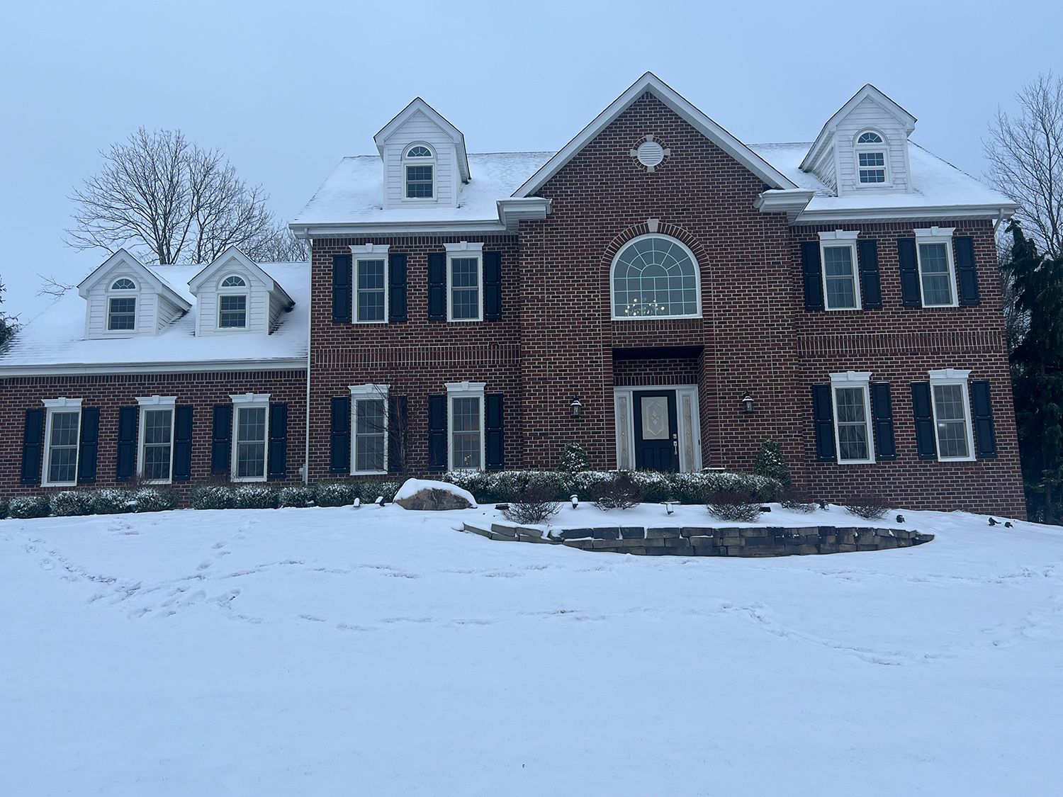 Two-story brick house covered in snow, with dark shutters, and three dormers under a cloudy sky.
