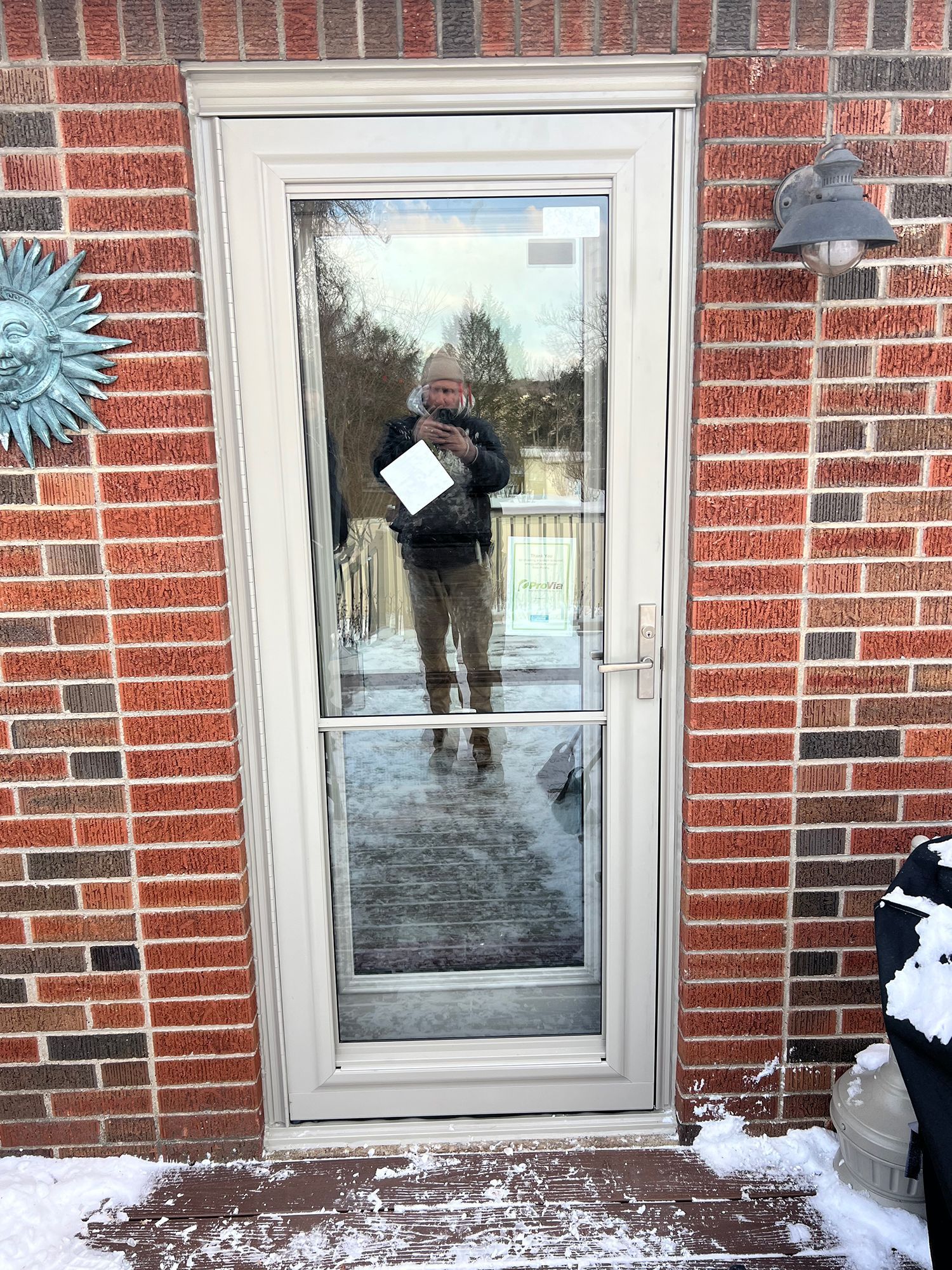 A person stands behind a clear glass door, winter scene outside. Snow on the deck and brick exterior.
