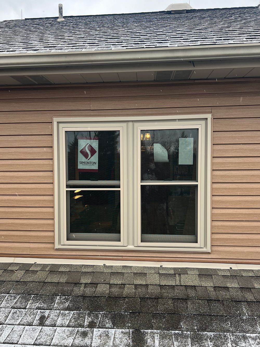 Window framed in beige against brown siding, snow on roof and shingles.