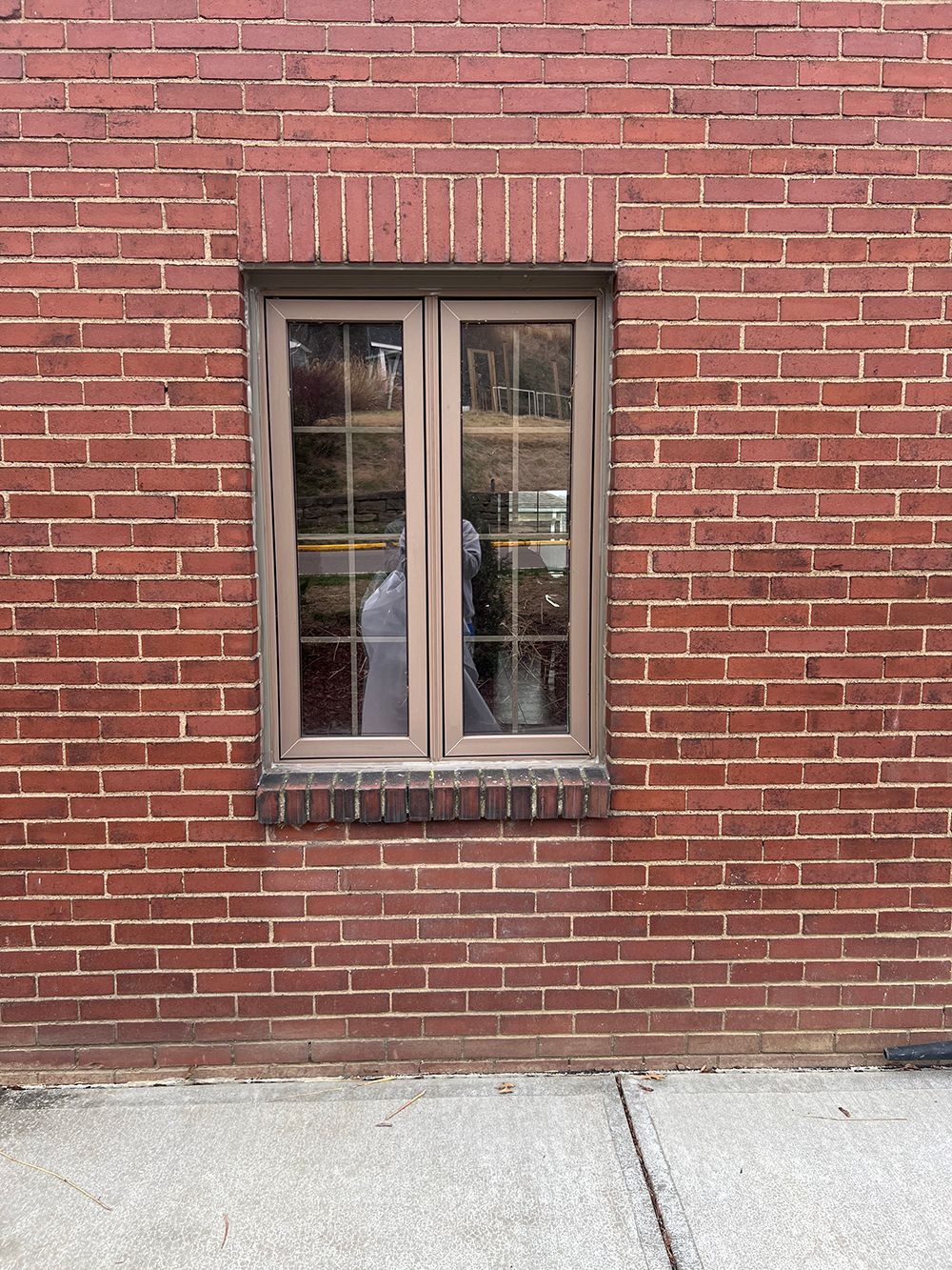 Brown window in a red brick wall with a concrete sidewalk.