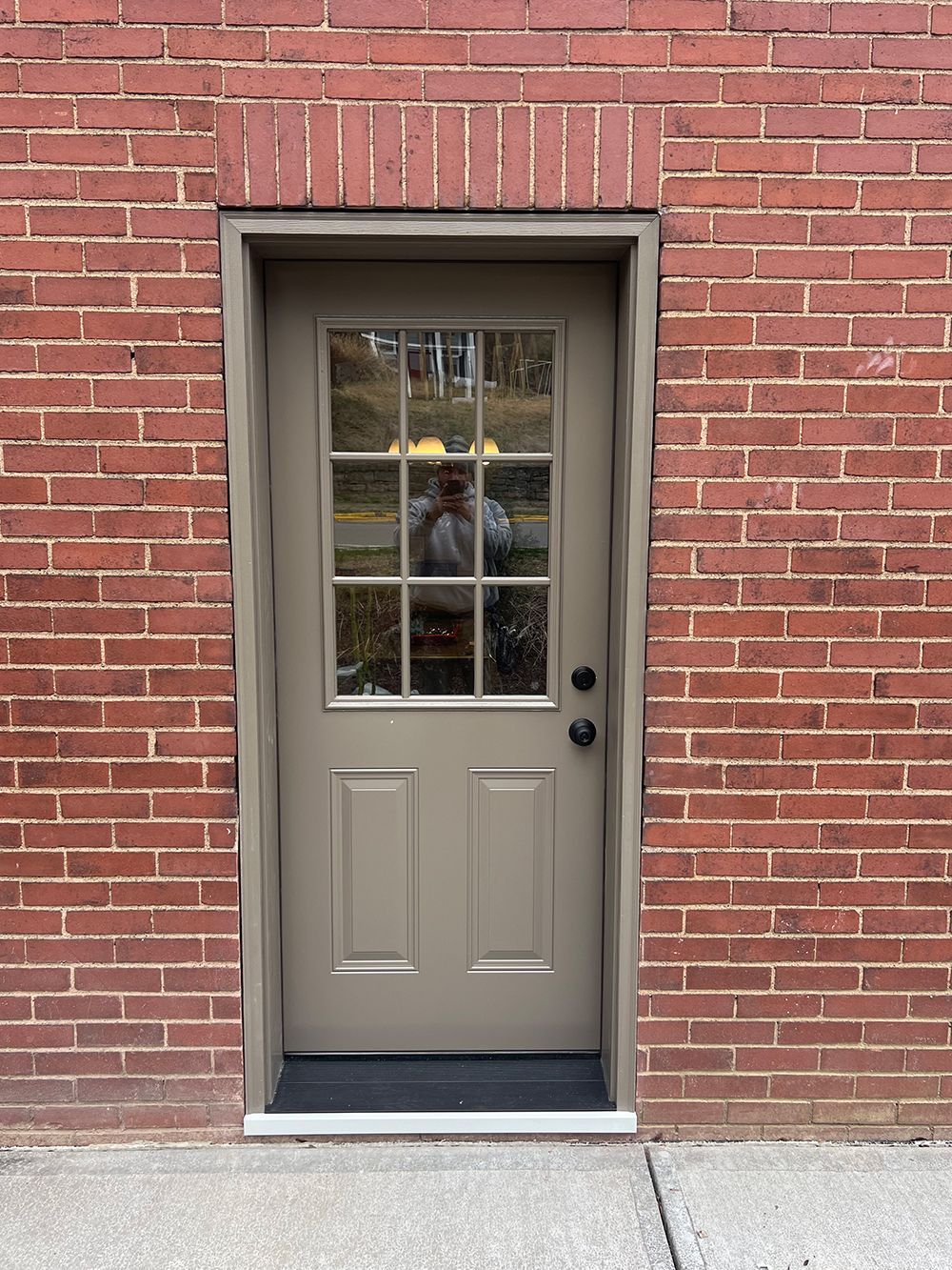 Tan door with glass panels set in a red brick wall. Person reflected in the glass.