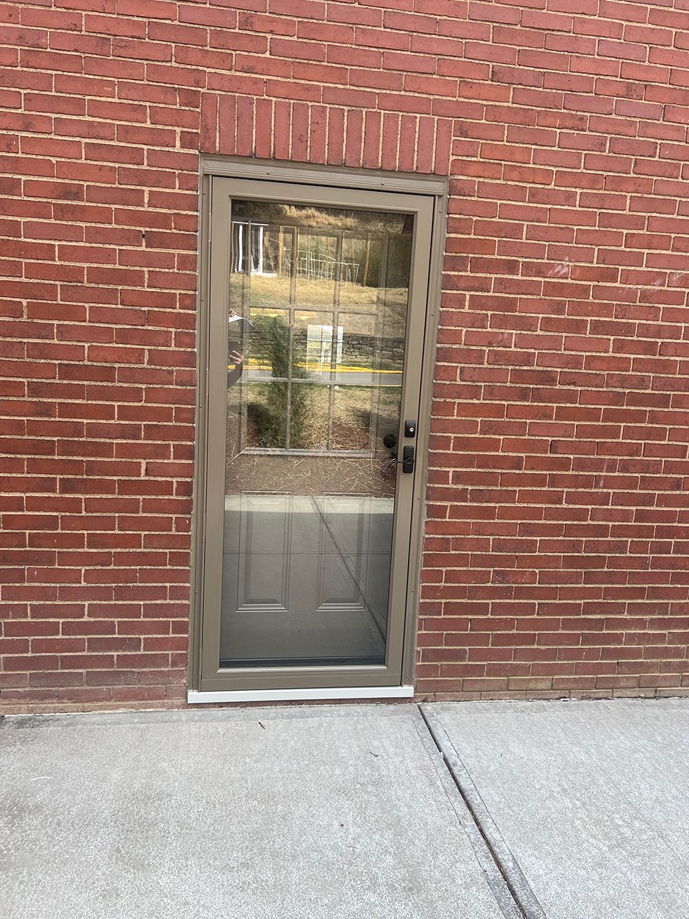 Tan-framed door set in a red brick wall, with a concrete floor. The door has a windowed upper section and solid lower.