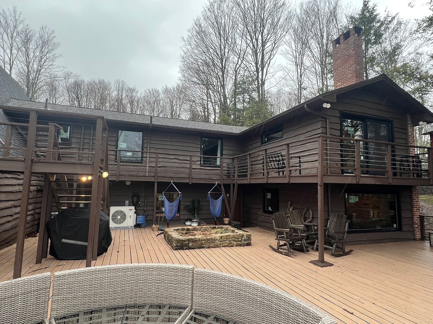 Brown cabin with a deck, fire pit, and chairs; surrounded by trees.