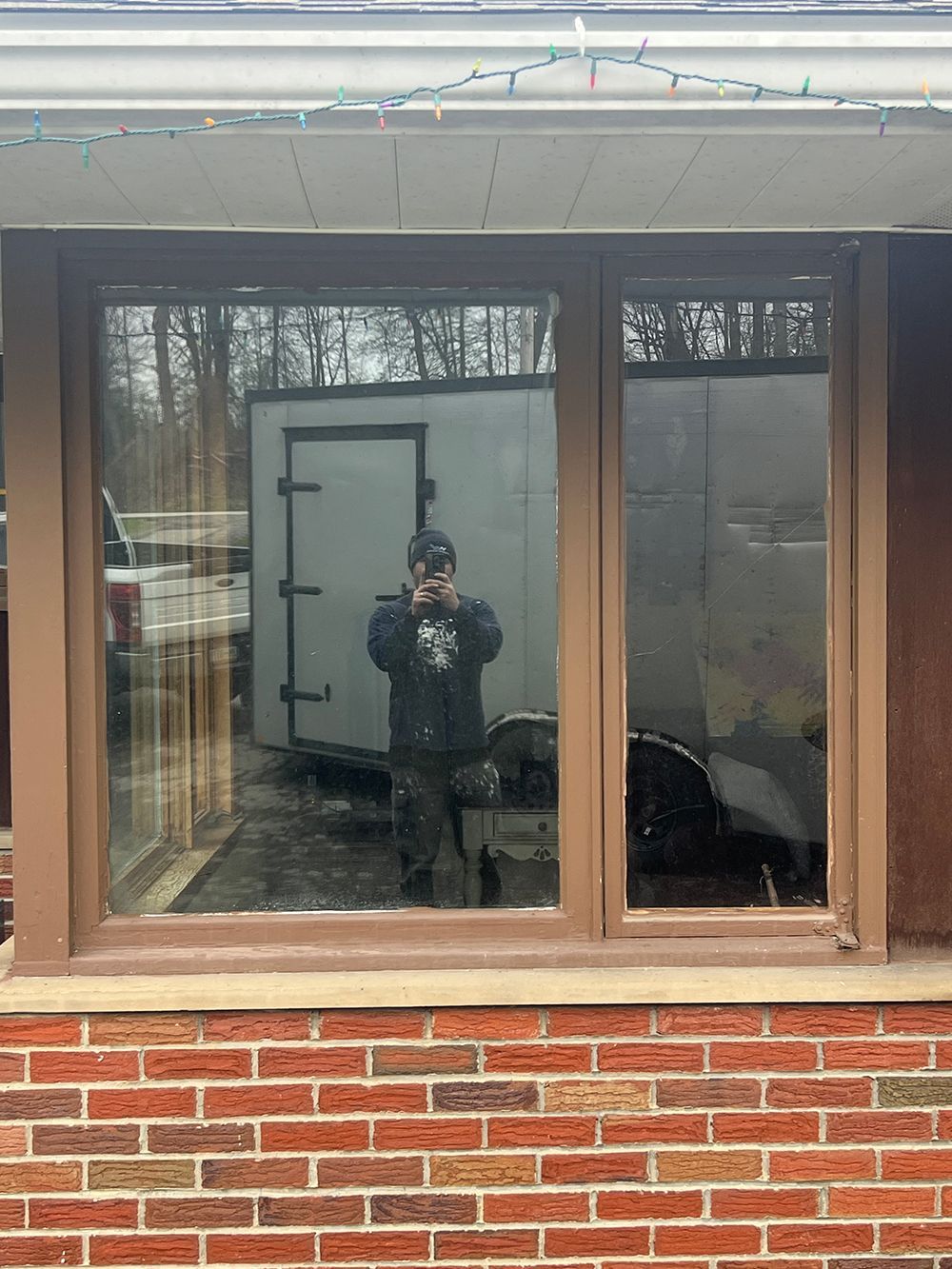 Brick building with brown-framed window reflecting person in dark jacket and a trailer.