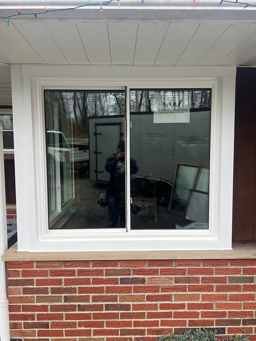 A white-framed sliding window set in a red brick wall, reflecting a person and outdoor objects.