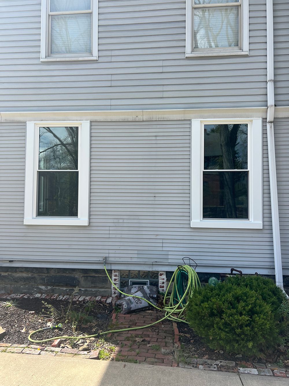 Exterior of a grey house with white-framed windows, a small bush, and a green hose in front.