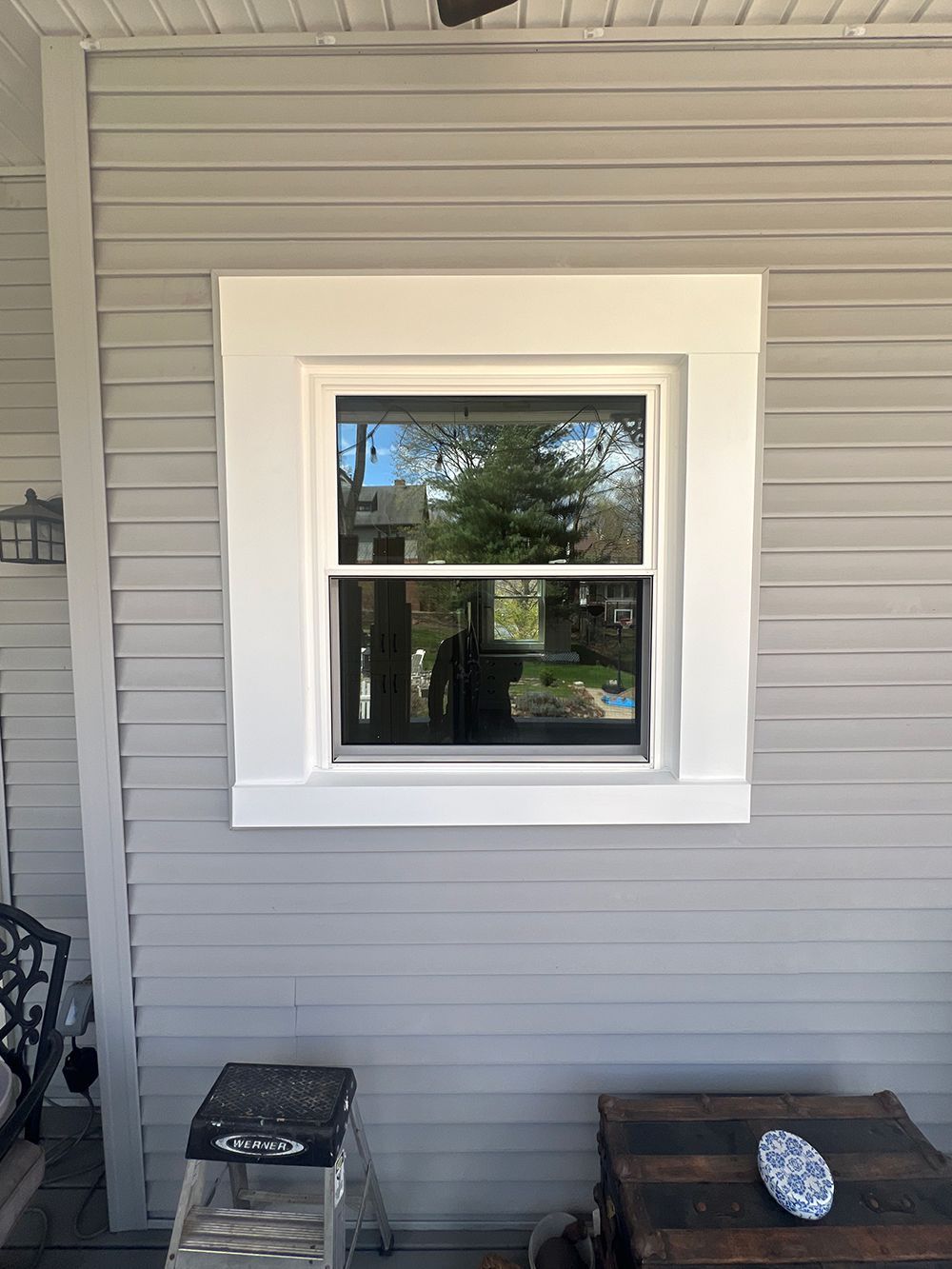 Window with white trim on gray siding; a tree is reflected in the glass.