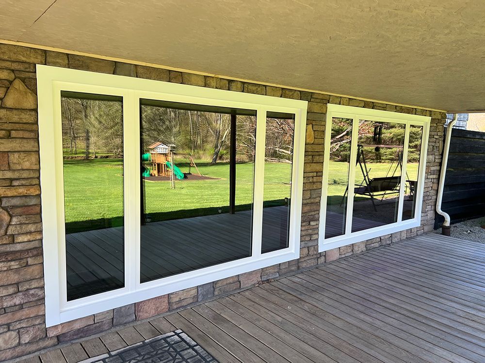 Two white-framed windows with reflective, dark glass on a stone and wooden porch, overlooking a yard.