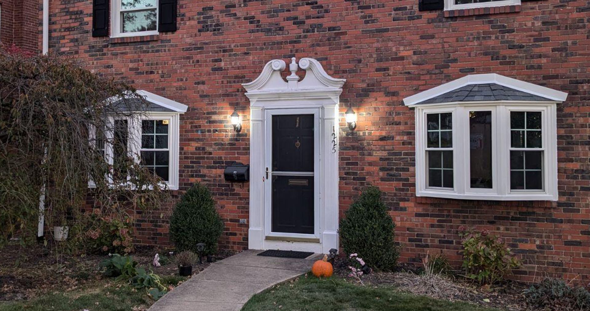 Red brick house with white door and bay windows, small walkway, and autumn décor.