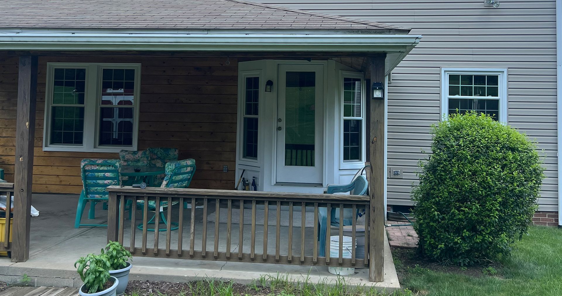Porch with wooden railing and trim, light blue chairs, and a white door, next to a green bush and light siding.