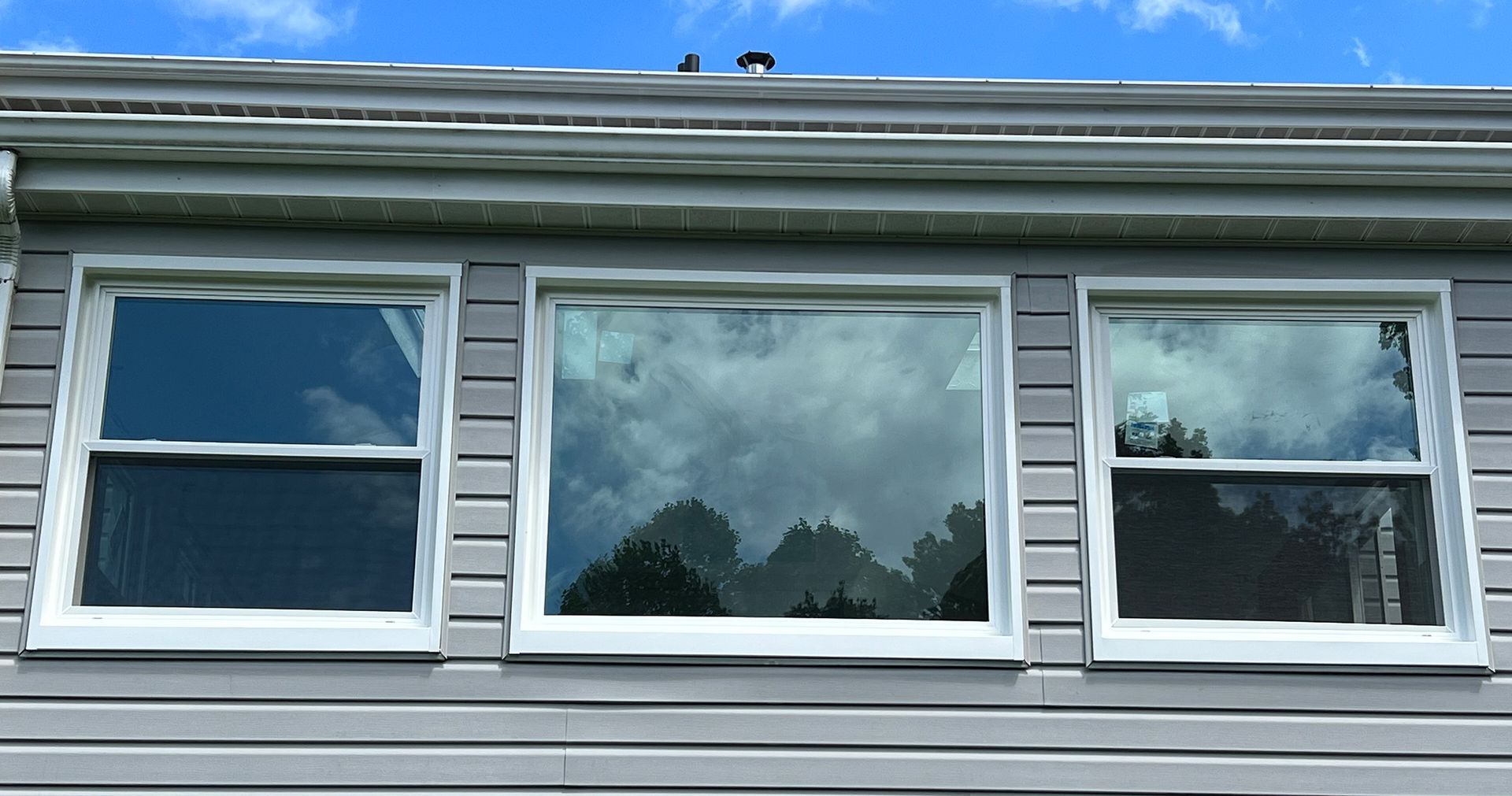 Three rectangular windows reflecting a cloudy sky and trees on a gray house exterior.